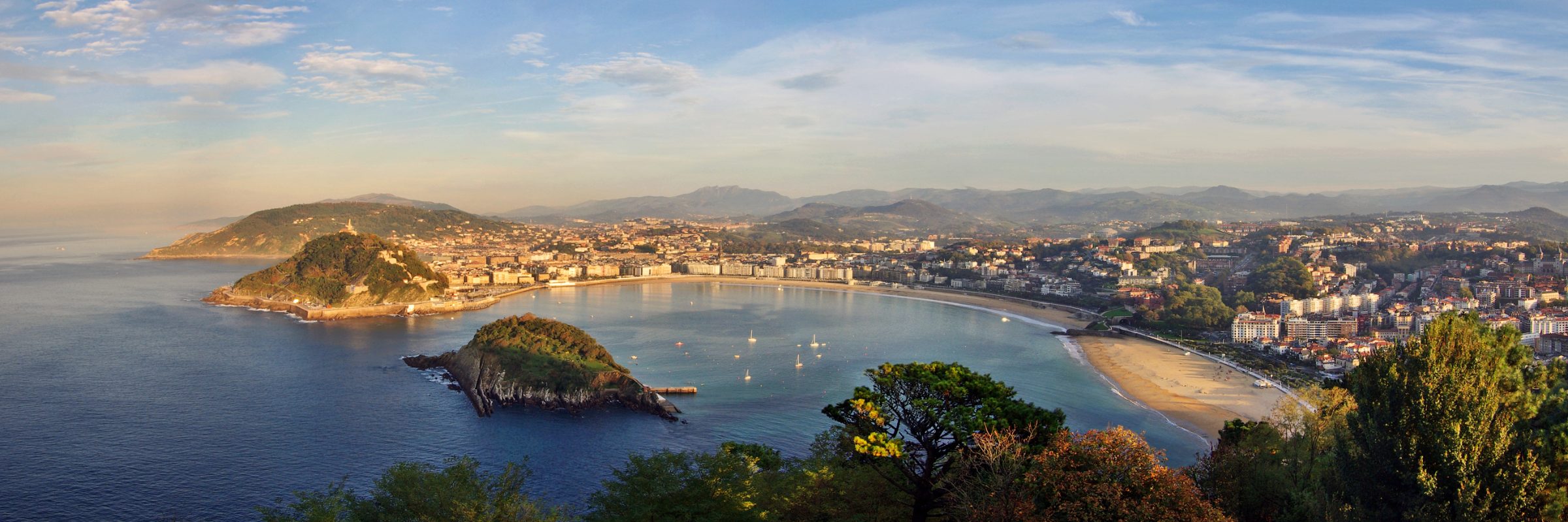 La Concha Bay in San Sebastián seen from Monte Igueldo in warm light, with Santa Clara island, Monte Urgull and the city curving around the beach.