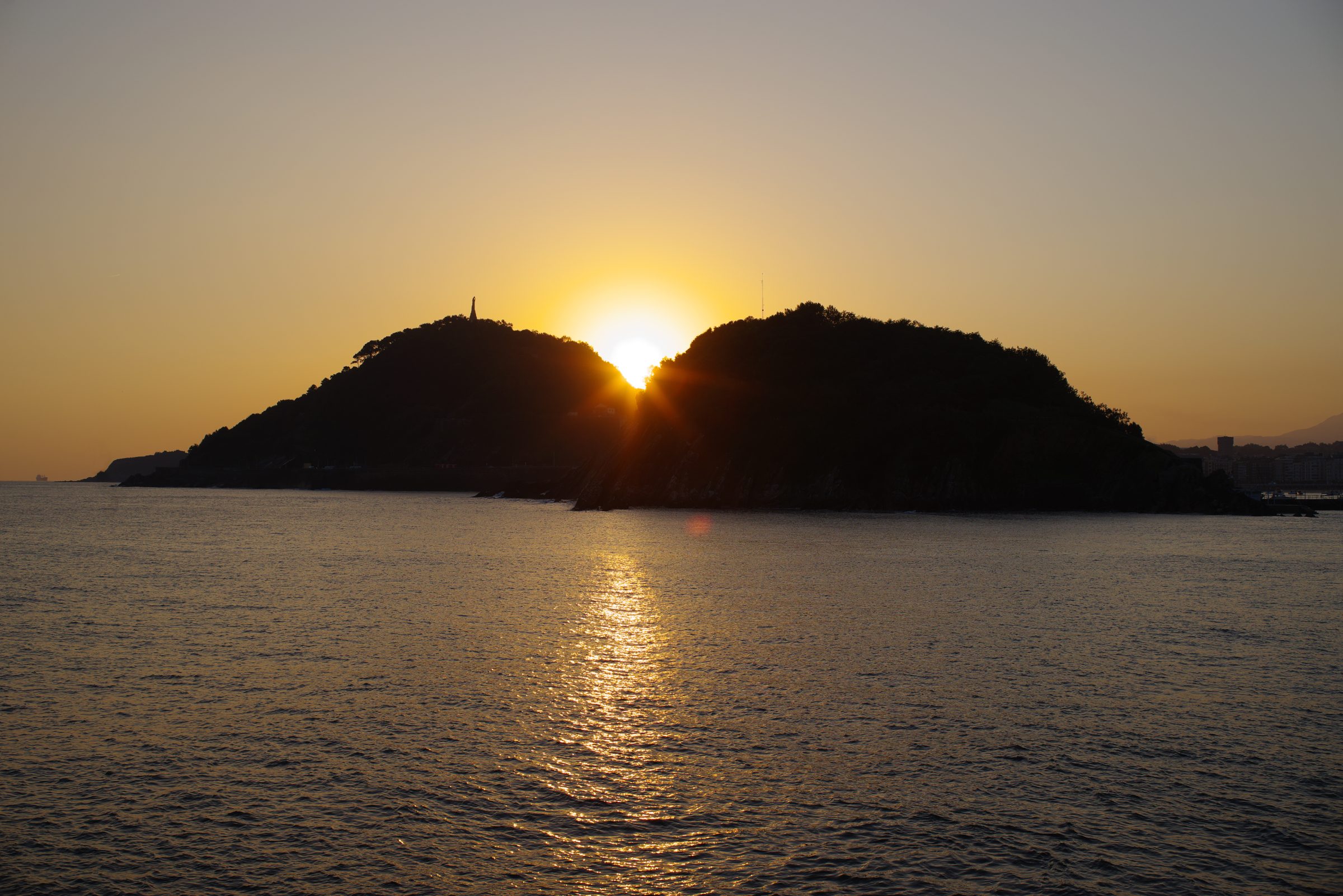 Sunrise between Monte Urgull and Santa Clara Island in San Sebastián, reflected on calm water.